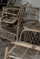 A collection of rustic, woven wicker chairs arranged on a wooden floor. The chairs have curved armrests and a woven back and seat design, giving a vintage and artisan feel.