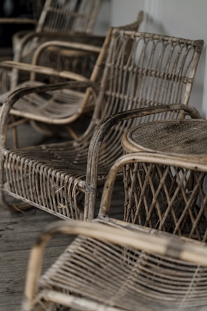 A collection of rustic, woven wicker chairs arranged on a wooden floor. The chairs have curved armrests and a woven back and seat design, giving a vintage and artisan feel.