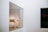 A modern art gallery with a large glass window revealing a room containing paintings on the wall. Inside, there are art installations or equipment on the floor, and the walls are minimalist and white.