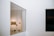 A modern art gallery with a large glass window revealing a room containing paintings on the wall. Inside, there are art installations or equipment on the floor, and the walls are minimalist and white.