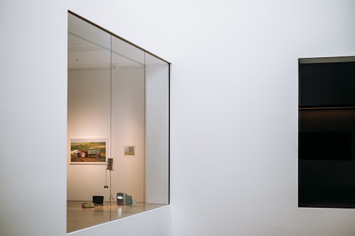 A modern art gallery with a large glass window revealing a room containing paintings on the wall. Inside, there are art installations or equipment on the floor, and the walls are minimalist and white.