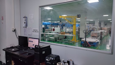 The image shows an industrial setting with a view through a large window. There is a control desk with computer monitors and various devices in the foreground. In the background, a factory floor with machinery, including a yellow robotic arm and various equipment, is visible. The space appears clean and organized, with box pallets stacked neatly.