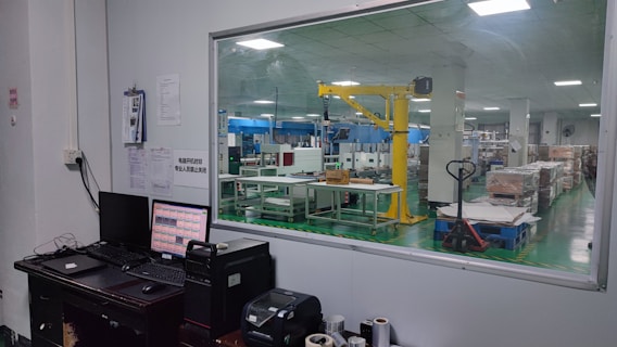 The image shows an industrial setting with a view through a large window. There is a control desk with computer monitors and various devices in the foreground. In the background, a factory floor with machinery, including a yellow robotic arm and various equipment, is visible. The space appears clean and organized, with box pallets stacked neatly.