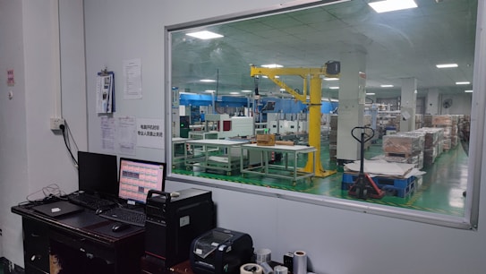 The image shows an industrial setting with a view through a large window. There is a control desk with computer monitors and various devices in the foreground. In the background, a factory floor with machinery, including a yellow robotic arm and various equipment, is visible. The space appears clean and organized, with box pallets stacked neatly.