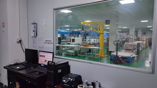 The image shows an industrial setting with a view through a large window. There is a control desk with computer monitors and various devices in the foreground. In the background, a factory floor with machinery, including a yellow robotic arm and various equipment, is visible. The space appears clean and organized, with box pallets stacked neatly.