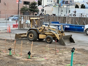 A construction site with a yellow backhoe loader parked on a dirt area. The surroundings include scattered sticks with markers, a wooden fence separating the site from a street, and a blue dumpster nearby. Various buildings and street signs are visible in the background.