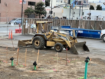 A construction site with a yellow backhoe loader parked on a dirt area. The surroundings include scattered sticks with markers, a wooden fence separating the site from a street, and a blue dumpster nearby. Various buildings and street signs are visible in the background.