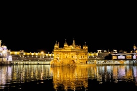 A beautifully illuminated temple is reflected in the calm waters of a surrounding pool, with intricate architecture and golden lighting contrasting against the dark night sky.