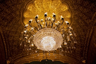 An ornate chandelier hangs from a richly decorated, golden ceiling. The chandelier features numerous crystal elements that catch and reflect the warm lighting, enhancing the opulent ambiance.