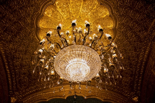 An ornate chandelier hangs from a richly decorated, golden ceiling. The chandelier features numerous crystal elements that catch and reflect the warm lighting, enhancing the opulent ambiance.