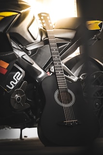 A black acoustic guitar is leaning against a sleek motorcycle, which features sharp lines and vibrant yellow accents, set against a warm backlit background.