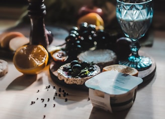A table spread with a variety of foods including sliced citrus fruits, a glass with a blue tint, a yogurt container, and a wooden board holding cheese and dark grapes. The scene is lit with natural light, casting soft shadows across the table.