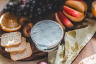 A close-up of a wooden board featuring assorted snacks. There are slices of orange, grapes, crackers, peach halves, and a sealed container labeled as blue cheese creme. A decorative napkin with a floral pattern is also present.