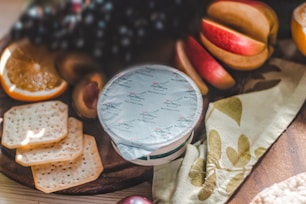 A close-up of a wooden board featuring assorted snacks. There are slices of orange, grapes, crackers, peach halves, and a sealed container labeled as blue cheese creme. A decorative napkin with a floral pattern is also present.