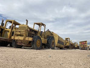 A row of large, heavy-duty construction vehicles is parked on a sandy terrain under a cloudy sky. The machinery appears to be aged and weathered, with visible rust and dirt. The vehicles are lined up in a slightly staggered formation, extending into the distance.