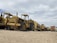 A row of large, heavy-duty construction vehicles is parked on a sandy terrain under a cloudy sky. The machinery appears to be aged and weathered, with visible rust and dirt. The vehicles are lined up in a slightly staggered formation, extending into the distance.