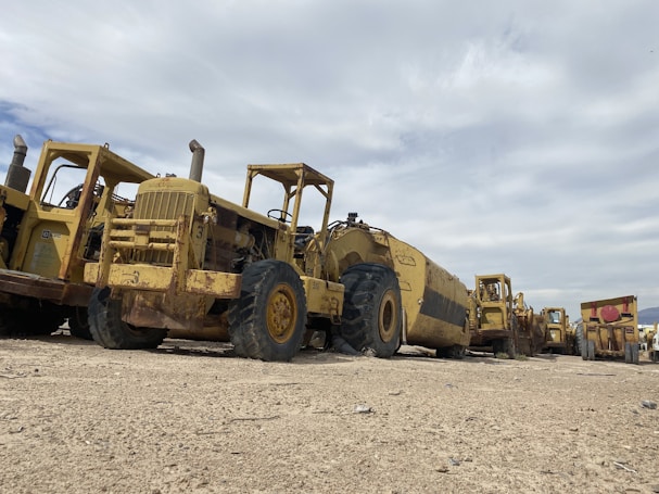 A row of large, heavy-duty construction vehicles is parked on a sandy terrain under a cloudy sky. The machinery appears to be aged and weathered, with visible rust and dirt. The vehicles are lined up in a slightly staggered formation, extending into the distance.