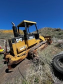 A yellow bulldozer is parked on uneven, grassy terrain next to a black tire. The vehicle has tracks for traversing difficult landscapes and a large front blade for moving earth. The background features a clear blue sky and rolling hills with patches of green and yellow vegetation.
