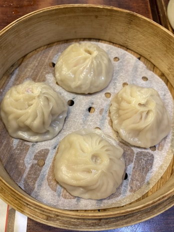 Four steamed dumplings with delicately pleated edges sit inside a round bamboo steamer basket. The dumplings have a slightly glossy texture and are resting on a perforated parchment paper.