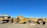 A collection of heavy machinery, including excavators and bulldozers, arranged on a gravel lot under a clear blue sky. The machines are predominantly yellow and have visible brand markings.