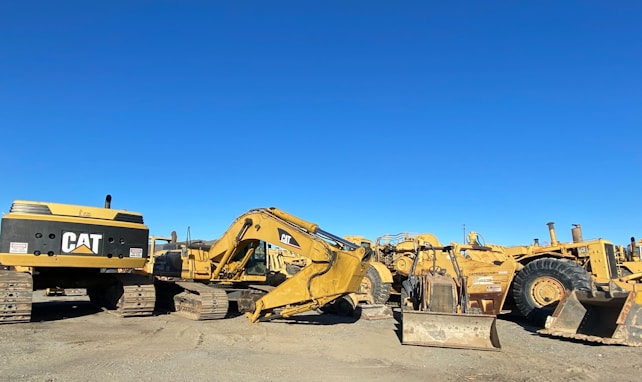 A collection of heavy machinery, including excavators and bulldozers, arranged on a gravel lot under a clear blue sky. The machines are predominantly yellow and have visible brand markings.