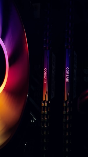 Two vertical Corsair RAM sticks with RGB lighting to the right of a colorful cooling fan in a computer setup. The fan displays a gradient of colors ranging from purple to red. The overall atmosphere is visually striking, emphasizing technology and modern design.