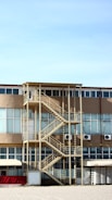 A multi-story building with a prominent external staircase. The structure features large windows and beige-colored walls. Air conditioning units are mounted on the exterior. The staircase is metal with a yellowish hue, and there is a flat roof above it. The ground is covered in gravel and the sky is clear and blue.
