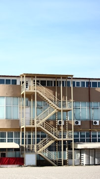 A multi-story building with a prominent external staircase. The structure features large windows and beige-colored walls. Air conditioning units are mounted on the exterior. The staircase is metal with a yellowish hue, and there is a flat roof above it. The ground is covered in gravel and the sky is clear and blue.