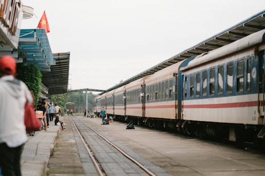 A train is stationed at a platform with people standing and walking around it. There is a Vietnamese flag on a building to the left and the platform is covered partially by a metal roof. The train tracks run alongside the platform, with greenery visible in the background.