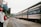 A train is stationed at a platform with people standing and walking around it. There is a Vietnamese flag on a building to the left and the platform is covered partially by a metal roof. The train tracks run alongside the platform, with greenery visible in the background.