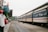 A train is stationed at a platform with people standing and walking around it. There is a Vietnamese flag on a building to the left and the platform is covered partially by a metal roof. The train tracks run alongside the platform, with greenery visible in the background.