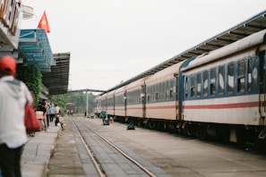 A train is stationed at a platform with people standing and walking around it. There is a Vietnamese flag on a building to the left and the platform is covered partially by a metal roof. The train tracks run alongside the platform, with greenery visible in the background.