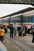 A group of people is on a train platform beside a stationary train. A conductor in uniform walks past several passengers, one of whom carries a suitcase. The scene suggests activity and travel, with people moving casually along the platform.