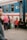 A group of people stands near a train platform, next to a blue and yellow train with open doors. A child in a red hoodie with the word 'PLAY' on it is facing the camera, while an older woman and some adults look on. The scene depicts a moment of transit or waiting.