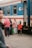 A group of people stands near a train platform, next to a blue and yellow train with open doors. A child in a red hoodie with the word 'PLAY' on it is facing the camera, while an older woman and some adults look on. The scene depicts a moment of transit or waiting.