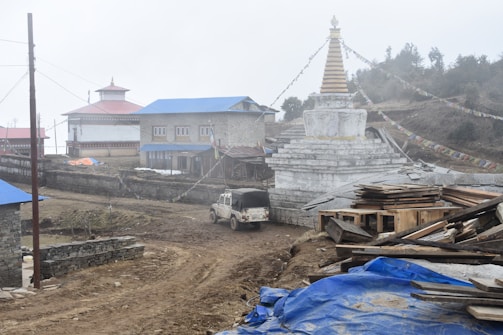 A rural scene featuring a stupa adorned with prayer flags, a few buildings with blue rooftops, and a dirt road with a parked SUV. There are some scattered construction materials covered by a blue tarp, and the atmosphere appears misty.