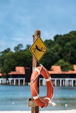 A wooden post featuring a yellow caution sign with an image of a person being pulled into water, likely indicating a water hazard. Attached below the sign is an orange lifebuoy secured with a white rope. In the background, there is a sandy beach, calm blue sea, and a structure with orange-tiled roofs surrounded by lush green trees.