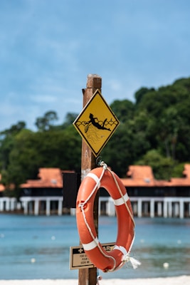 A wooden post featuring a yellow caution sign with an image of a person being pulled into water, likely indicating a water hazard. Attached below the sign is an orange lifebuoy secured with a white rope. In the background, there is a sandy beach, calm blue sea, and a structure with orange-tiled roofs surrounded by lush green trees.