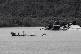 A partially submerged shipwreck is visible in a body of water, with a densely forested hillside in the background. To the right, an industrial structure, possibly a dredging operation, is present onshore with machinery and mounds of sand.