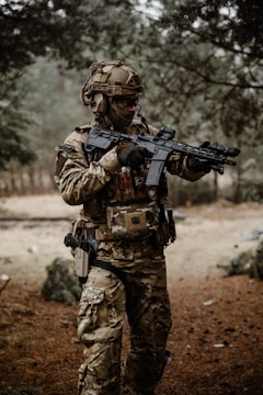A person dressed in full military tactical gear, including a helmet and camouflage uniform, stands in a wooded area. They are holding a rifle with both hands and appear to be in a ready stance, indicating alertness. The setting is natural with pine trees and a forest floor covered in brown pine needles.