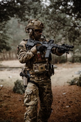 A person dressed in full military tactical gear, including a helmet and camouflage uniform, stands in a wooded area. They are holding a rifle with both hands and appear to be in a ready stance, indicating alertness. The setting is natural with pine trees and a forest floor covered in brown pine needles.