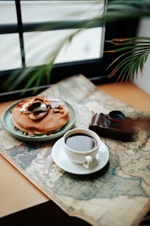 A cup of coffee sits on an antique map beside a plate of pancakes topped with slices of fruit and nuts. An old camera, partially in view, adds a vintage touch to the cozy setting. A leafy plant frames the composition, suggesting a peaceful and relaxed atmosphere. Sunlight filters through a window in the background.
