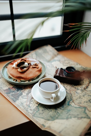 A cup of coffee sits on an antique map beside a plate of pancakes topped with slices of fruit and nuts. An old camera, partially in view, adds a vintage touch to the cozy setting. A leafy plant frames the composition, suggesting a peaceful and relaxed atmosphere. Sunlight filters through a window in the background.