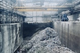A large industrial interior with heaps of shredded material piled in the center, surrounded by high concrete walls. Overhead, a yellow gantry crane runs along the ceiling with a large blue claw attachment, seemingly used for moving the material. The space has a metallic and utilitarian appearance, with steel girders and pipes visible.