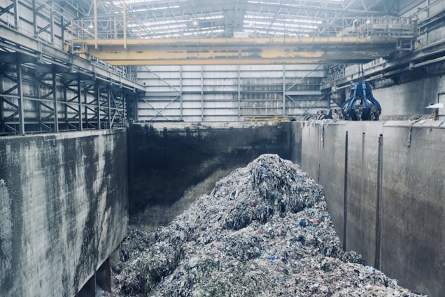 A large industrial interior with heaps of shredded material piled in the center, surrounded by high concrete walls. Overhead, a yellow gantry crane runs along the ceiling with a large blue claw attachment, seemingly used for moving the material. The space has a metallic and utilitarian appearance, with steel girders and pipes visible.