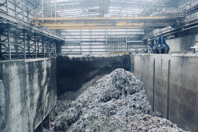 A large industrial interior with heaps of shredded material piled in the center, surrounded by high concrete walls. Overhead, a yellow gantry crane runs along the ceiling with a large blue claw attachment, seemingly used for moving the material. The space has a metallic and utilitarian appearance, with steel girders and pipes visible.
