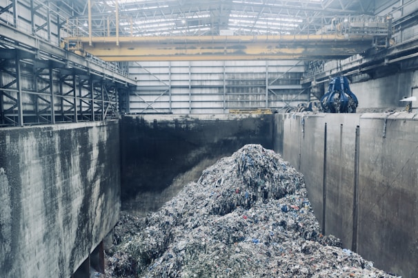 A large industrial interior with heaps of shredded material piled in the center, surrounded by high concrete walls. Overhead, a yellow gantry crane runs along the ceiling with a large blue claw attachment, seemingly used for moving the material. The space has a metallic and utilitarian appearance, with steel girders and pipes visible.