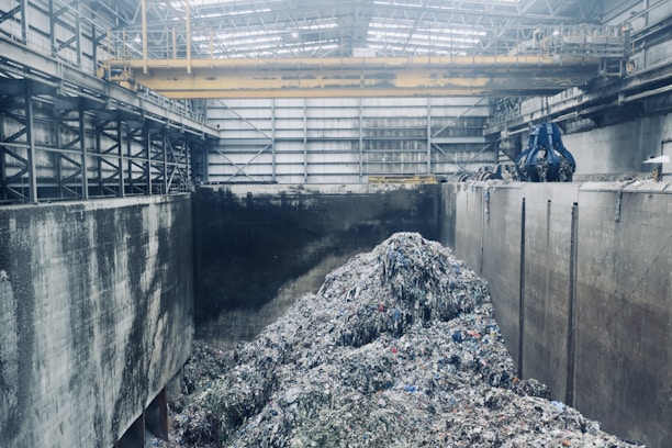 A large industrial interior with heaps of shredded material piled in the center, surrounded by high concrete walls. Overhead, a yellow gantry crane runs along the ceiling with a large blue claw attachment, seemingly used for moving the material. The space has a metallic and utilitarian appearance, with steel girders and pipes visible.