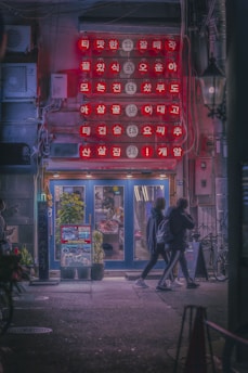 A dimly-lit street scene with a storefront displaying bright red signs featuring Asian characters. The entrance is flanked by potted plants and illuminated by soft light. Two people dressed in dark clothing walk past the store, adding a sense of motion to the calm atmosphere. Bicycles are parked nearby, and neon accents contribute to the urban night setting.