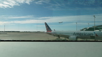 A view from behind glass panes with horizontal lines looking out onto an airport runway. A large airplane is parked on the tarmac with the word 'American' partially visible on the body. Another airplane is in the distance taking off against a backdrop of scattered clouds and blue sky.